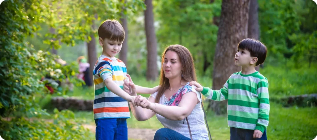 Young woman mother applying insect repellent to her two son before forest hike beautiful summer day